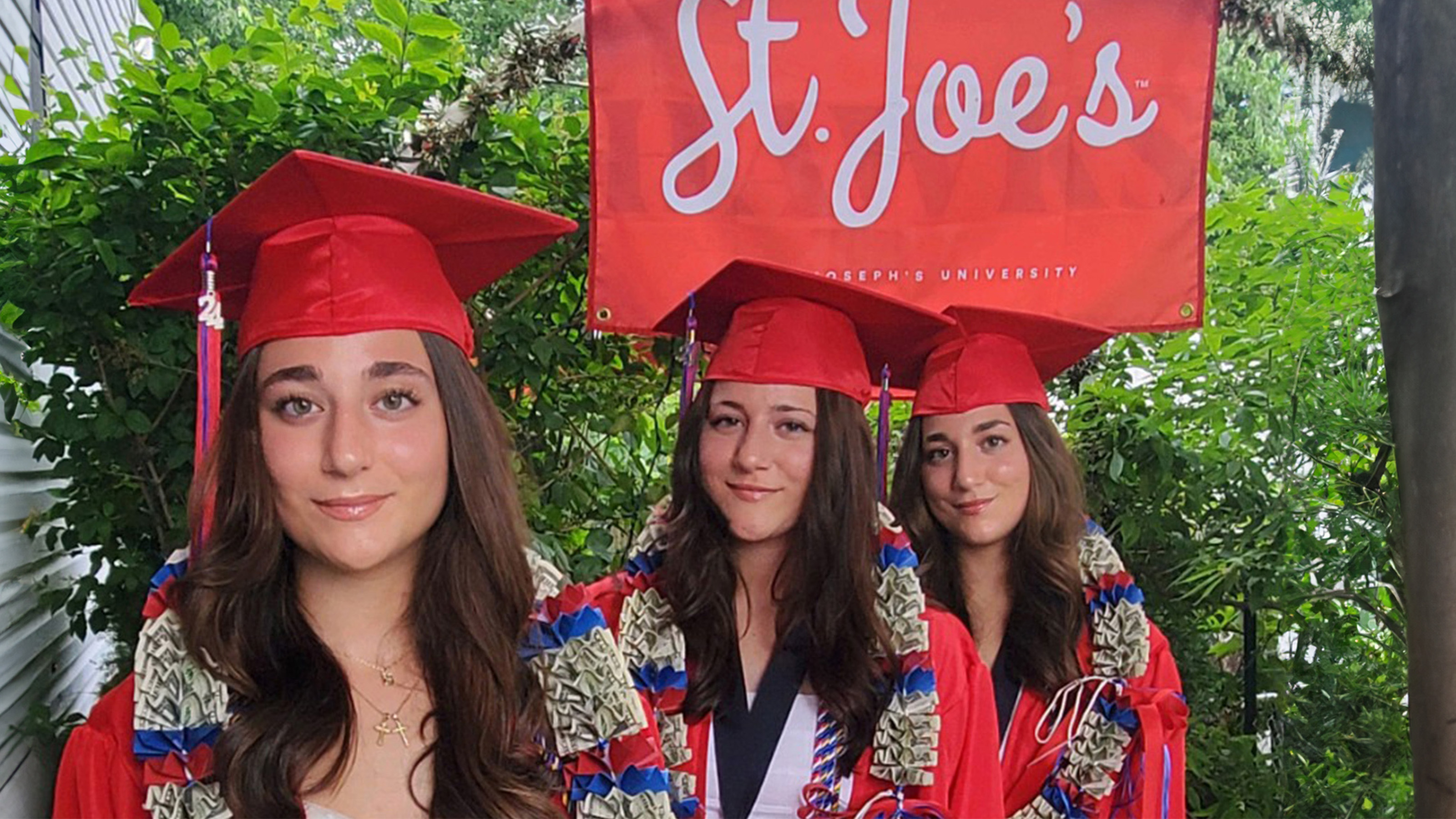 Oliveira sisters standing under a red St. Joe's flag in red caps and gowns