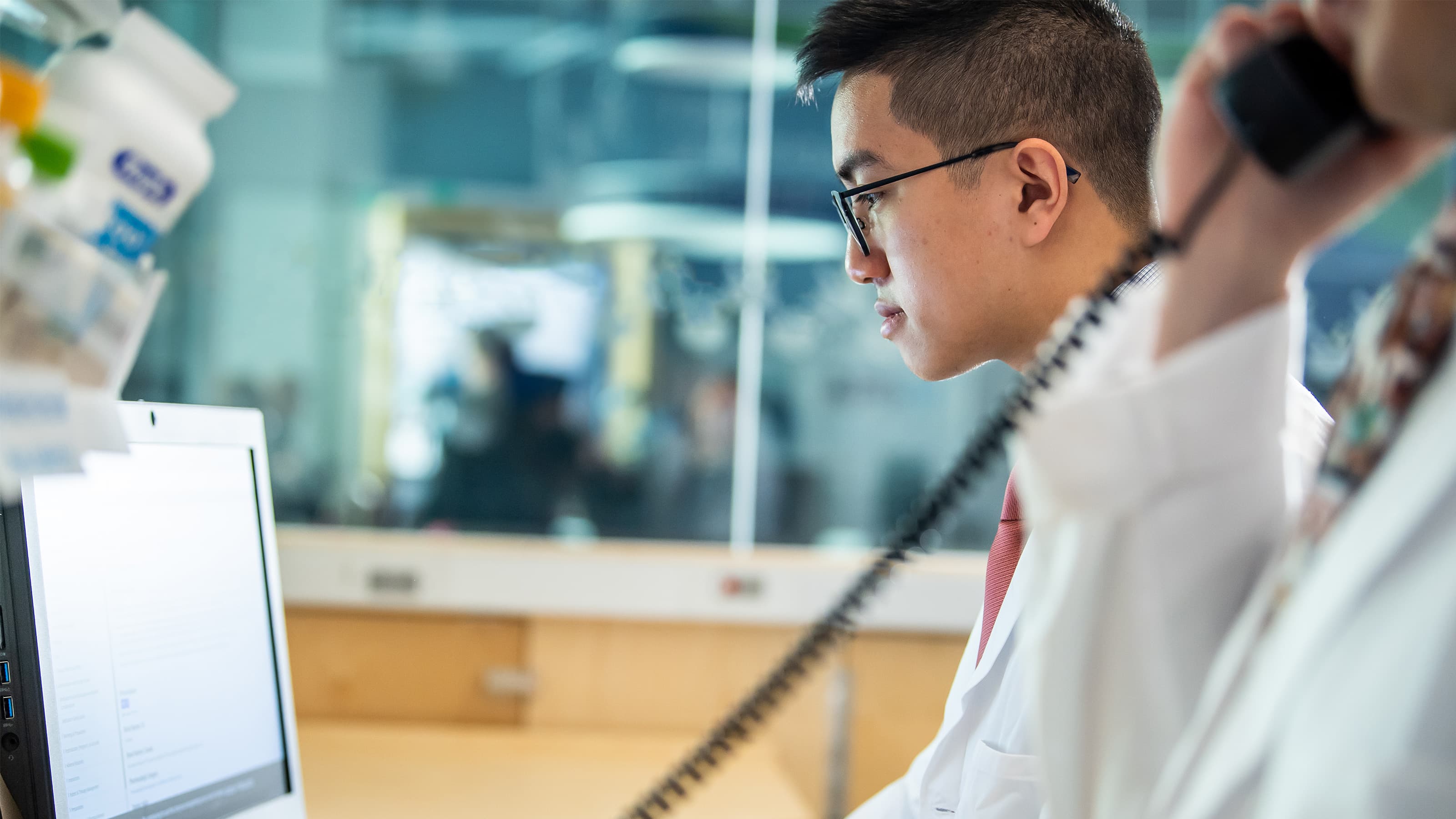 Male student in white coat typing at a computer in a pharmacy