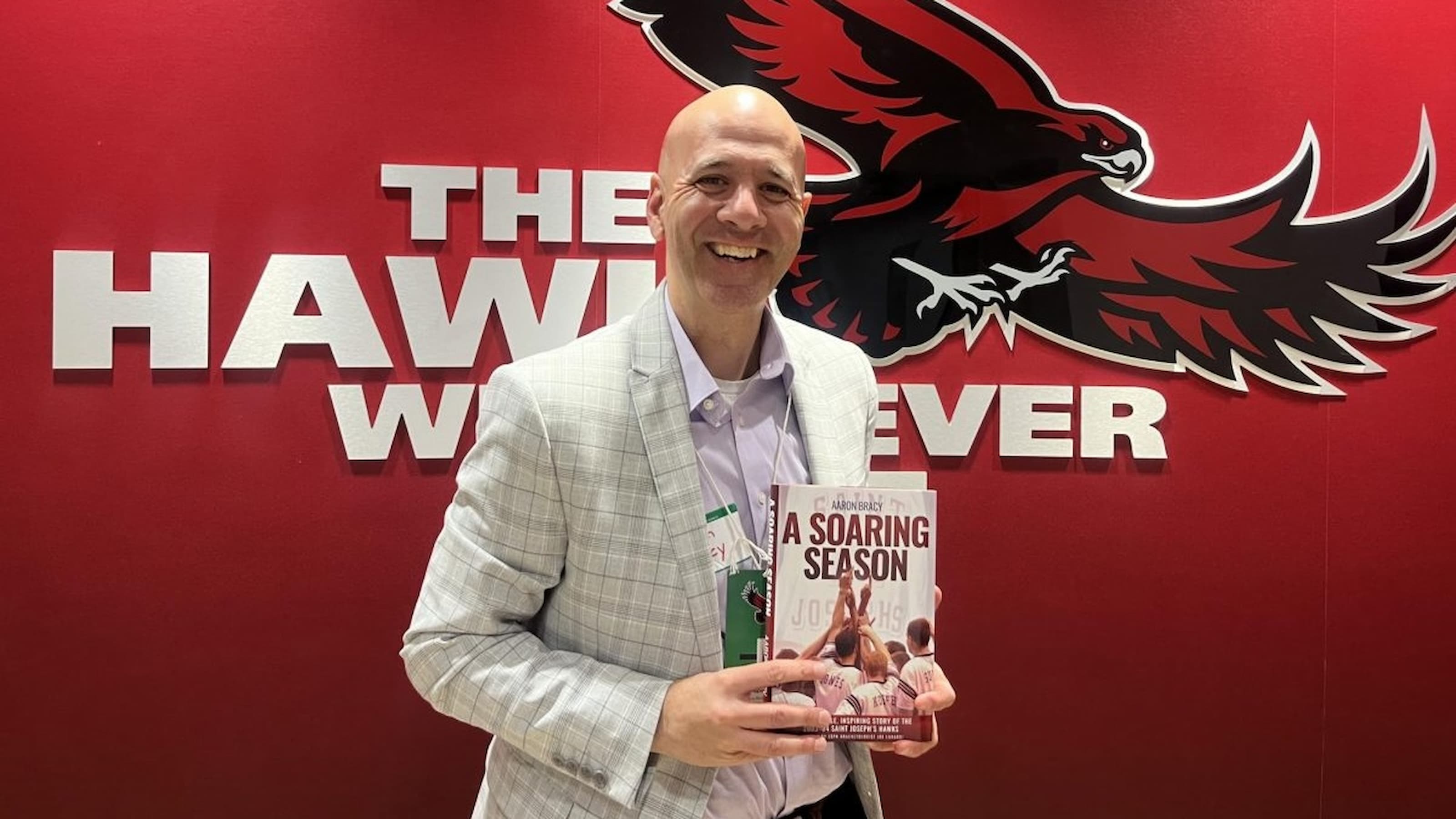 Aaron Bracy, BS ’98, MS ’99, poses with his book in front of the Hawk logo.