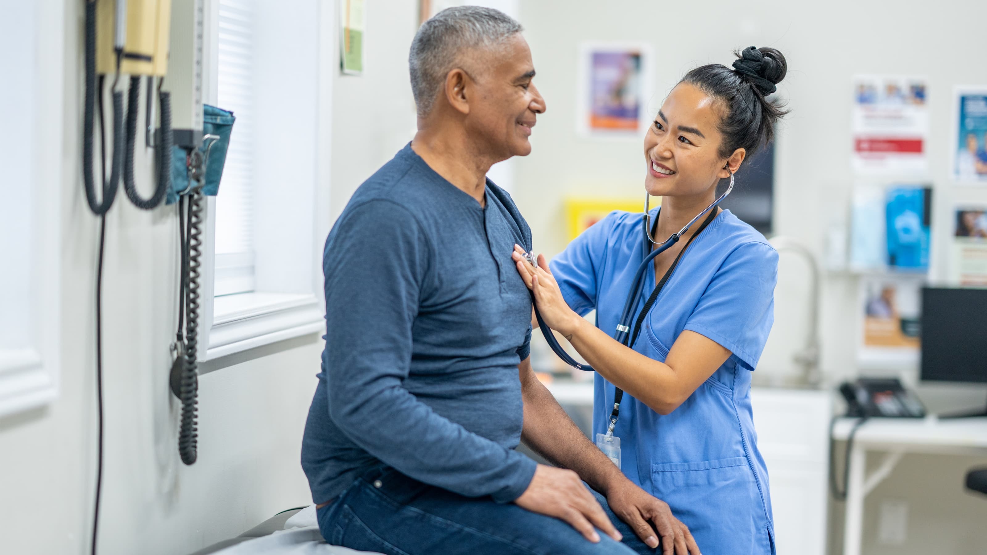 A nurse in blue scrubs listens to a patient's heart with a stethoscope.
