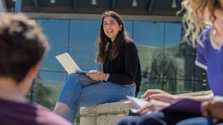 A smiling student sits outside Post Learning Commons with her laptop