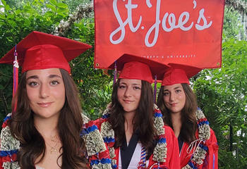 Oliveira sisters standing under a red St. Joe's flag in red caps and gowns