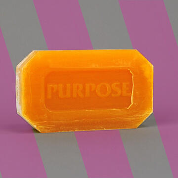 Amber bar of soap with "PURPOSE" imprinted on it placed against a backdrop of grey and pink diagonal stripes