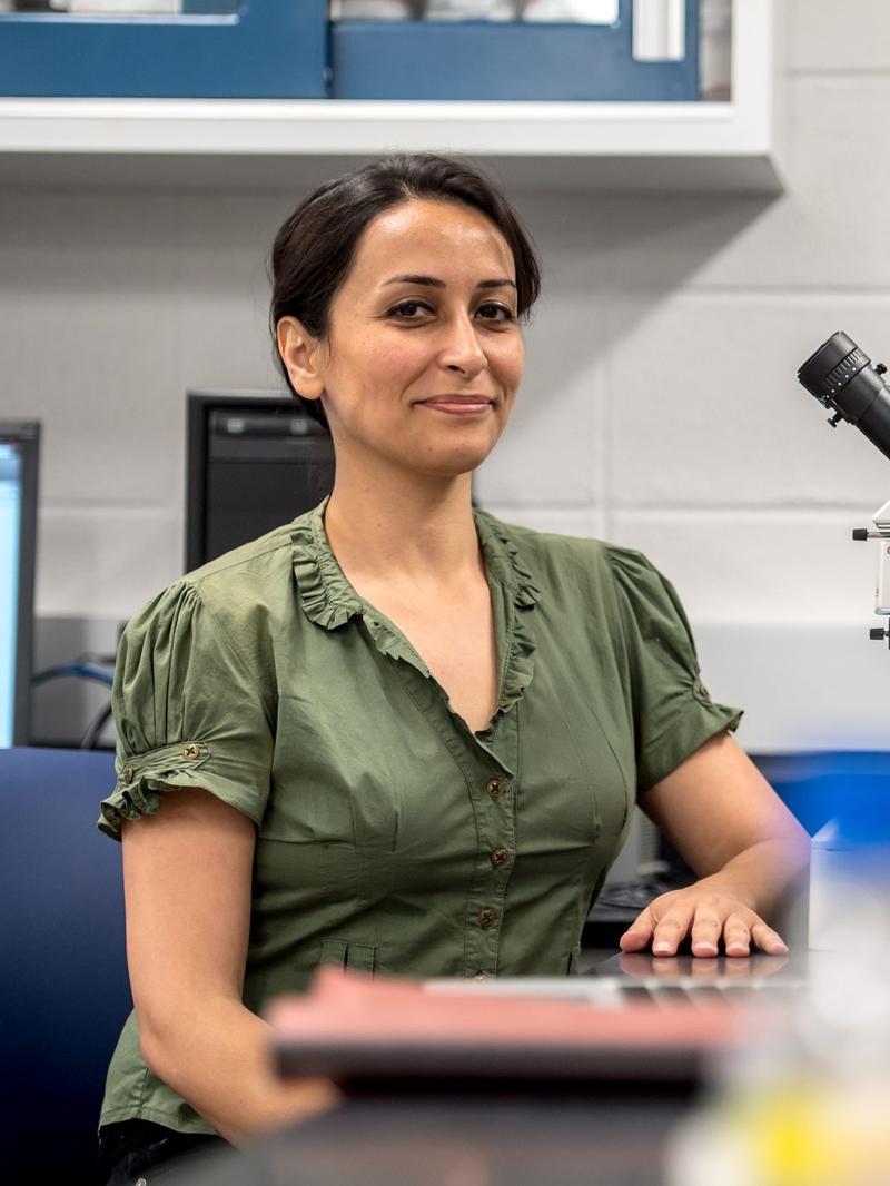 a woman wearing a green blouse and sitting inside a laboratory, surrounded with microscopes